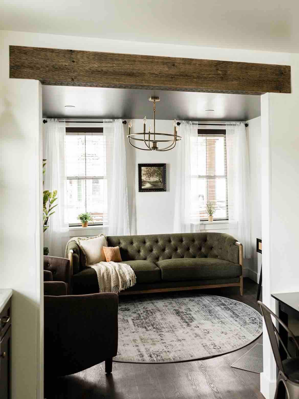 A cozy living room features a green tufted sofa accompanied by a dark armchair. Natural light filters through two windows adorned with sheer white curtains, while a round area rug anchors the space. A modest chandelier hangs from the ceiling, enhancing the room's warmth.