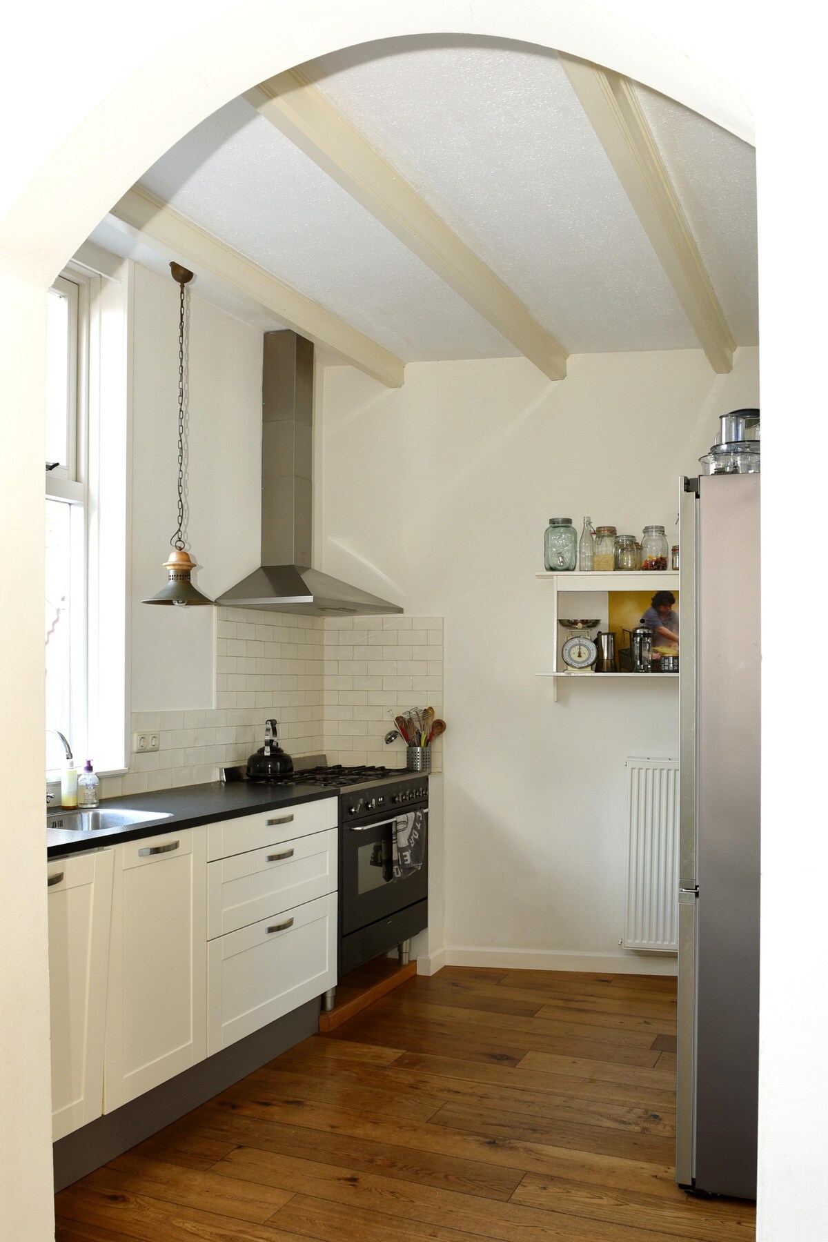 A modern kitchen features a blend of white cabinetry and dark countertops, complemented by stainless steel appliances. Natural light enters through a nearby window, highlighting the wooden flooring. An archway frames the kitchen, adding character to the inviting space.
