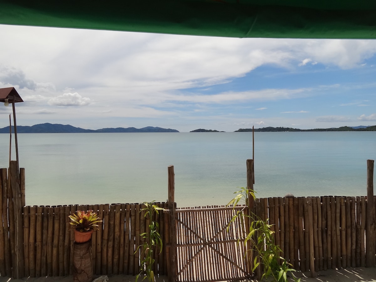 A serene seascape is displayed, showcasing calm waters meeting a clear sky. A bamboo fence borders the foreground, with potted plants adding greenery. Distant mountains are visible across the horizon, blending gently into the tranquil ocean, inviting a sense of peace.