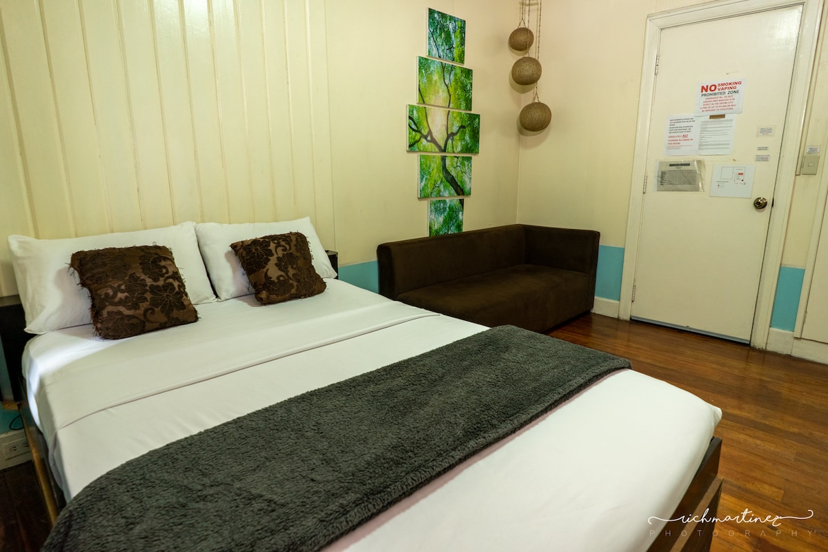 A well-appointed room features a spacious bed, adorned with a dark gray throw and accented by decorative pillows. A cozy seating area is positioned against the wall, with nature-themed artwork displayed above. Light wood flooring complements the soft color palette of the walls.