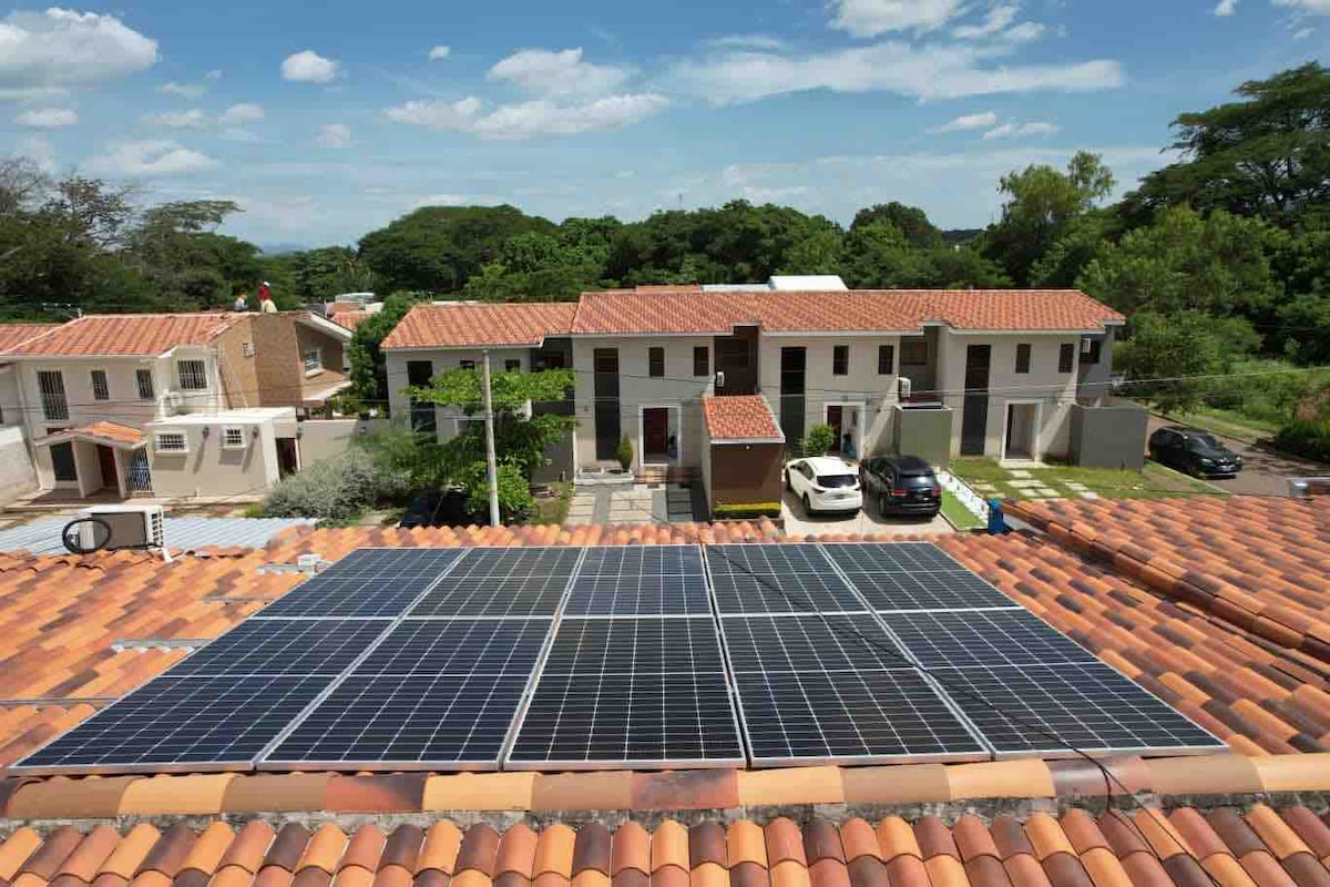 Solar panels are installed on the terracotta roof, reflecting a commitment to sustainability. In the background, residential buildings with red tile roofs are situated among landscaped green areas, creating a peaceful atmosphere in the private neighborhood.