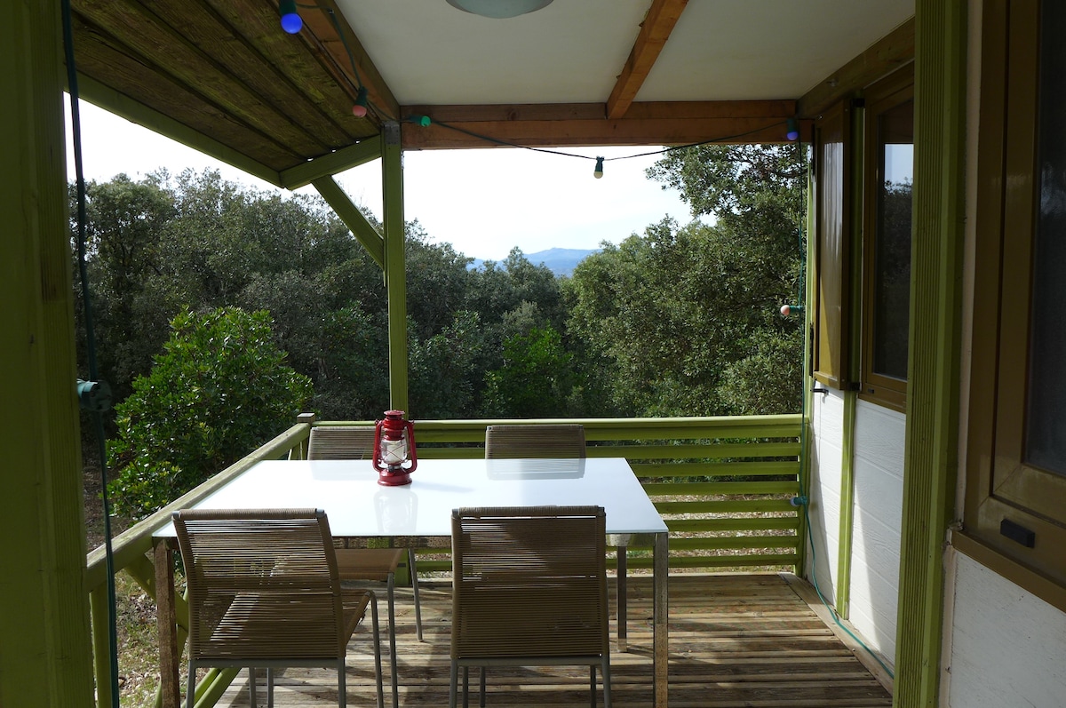A shaded outdoor seating area is visible, featuring a white table surrounded by four light brown chairs. A red lantern is positioned at the center of the table. Lush greenery is present in the background, with rolling hills suggested in the distance.
