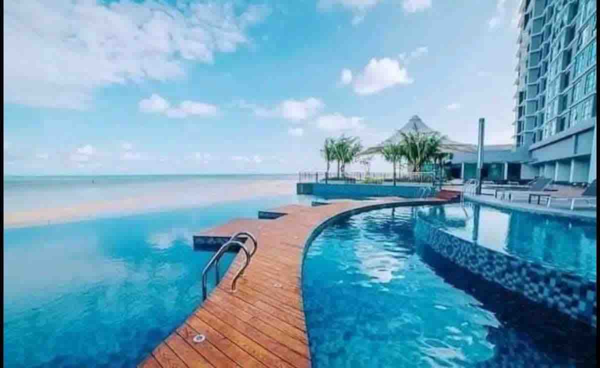 A tranquil outdoor swimming pool is set against a serene ocean backdrop. A wooden walkway meanders through the water, leading to a shaded seating area. Lush palm trees are visible, complementing the bright blue sky scattered with clouds.