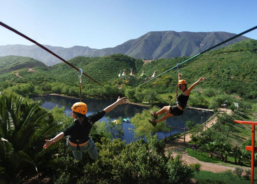 Two individuals are seen gliding on a zip line, suspended over a lush green landscape. Rolling hills stretch in the background, with clear blue skies above. A calm body of water is visible below, reflecting the surrounding greenery.