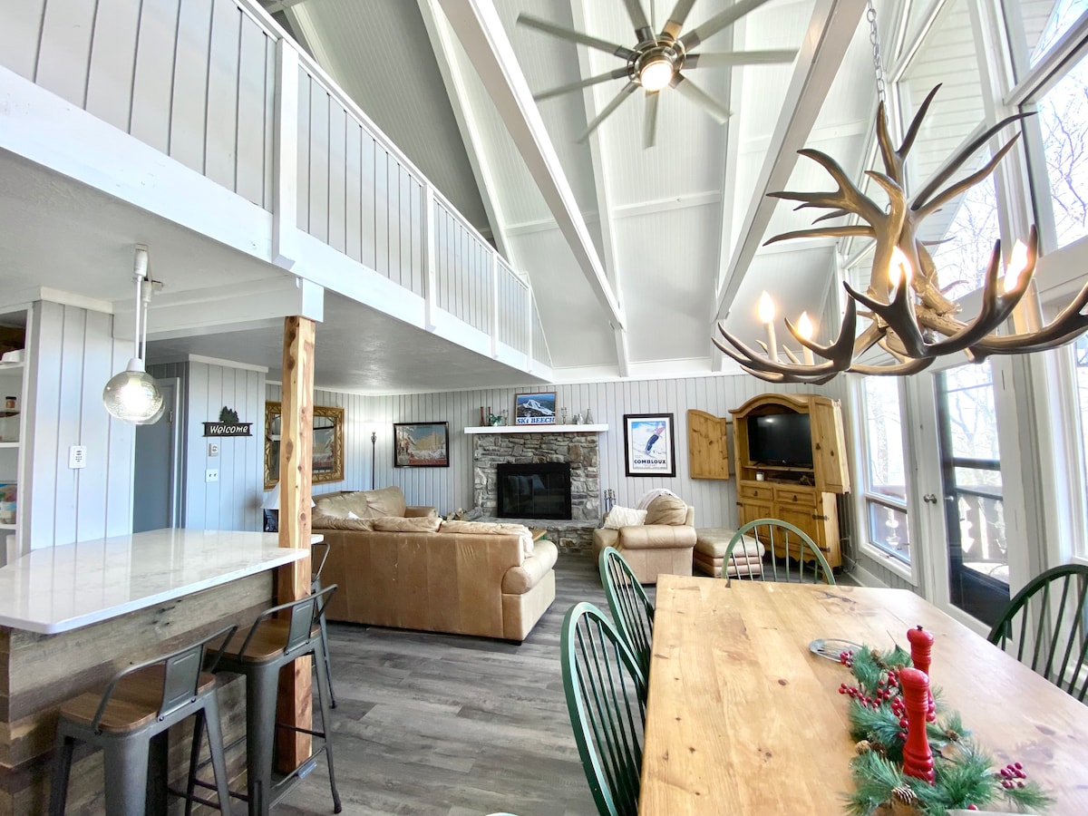 An open living space blends the kitchen and dining areas with a wooden dining table surrounded by green chairs. A stone fireplace anchors the room, with large windows providing natural light. The vaulted ceiling features a ceiling fan and wooden beams, enhancing the airy feel.