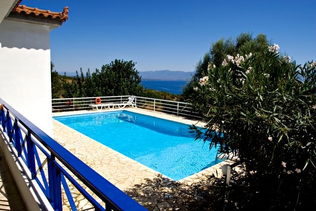 A private swimming pool is set amidst lush greenery, featuring a clear blue water surface. Surrounding the pool are sun loungers and a safety buoy, while views of distant mountains and the sea can be seen beyond the elegant stone terrace.