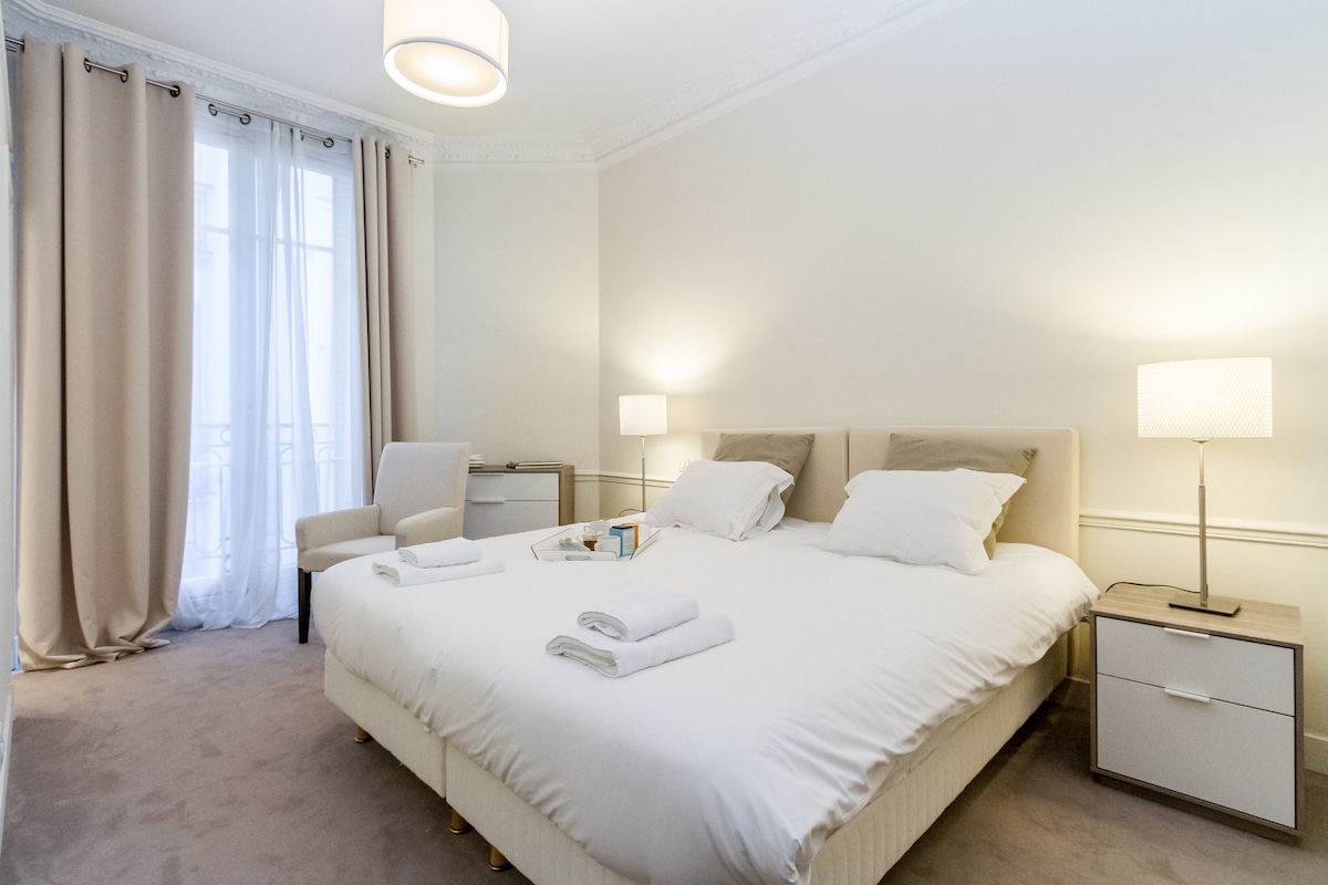 A bright bedroom features a spacious bed with neatly arranged pillows. Soft beige curtains frame a large window, allowing natural light to fill the room. Two bedside lamps offer gentle lighting, while a small chair and a nightstand complete the inviting space.