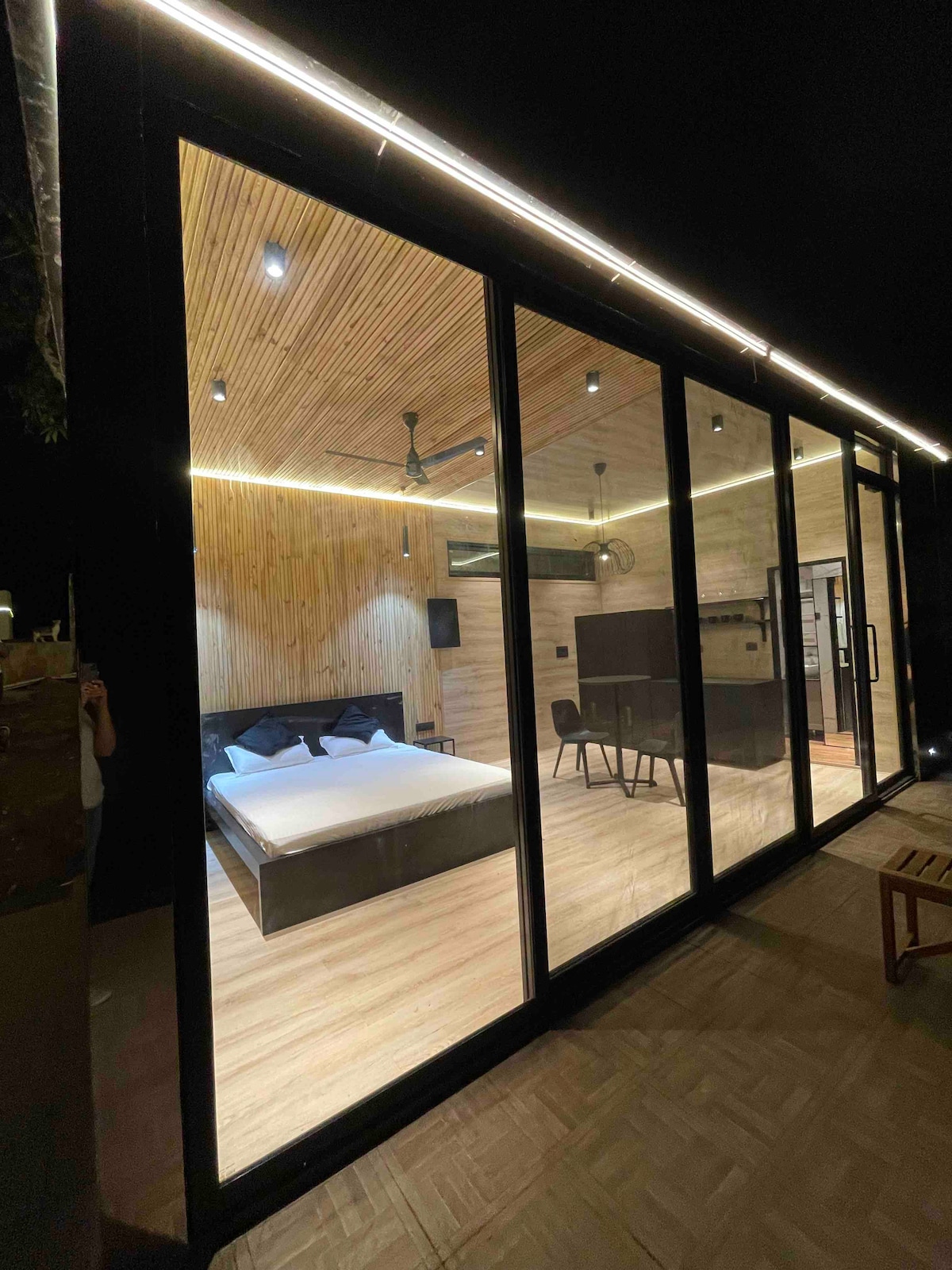 A modern cabin is visible through expansive glass doors, offering a view of the spacious interior. The wooden ceiling complements the minimalist design, while a large bed is prominently placed within the room. Soft lighting enhances the cozy atmosphere.
