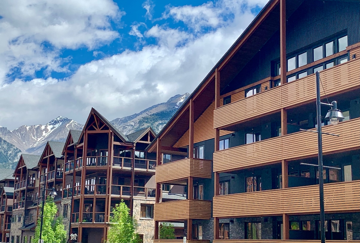 The exterior of the luxury resort is showcased, featuring a contemporary design with wood accents and expansive balconies. Flourishing green trees surround the entrance, and majestic mountains rise in the background under a partly cloudy sky.