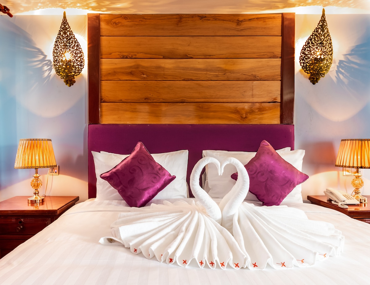 The spacious bed is adorned with a decorative headboard and features elegant purple pillows. White towels are arranged to form swans at the center, while two bedside lamps provide gentle illumination, enhancing the overall ambiance.