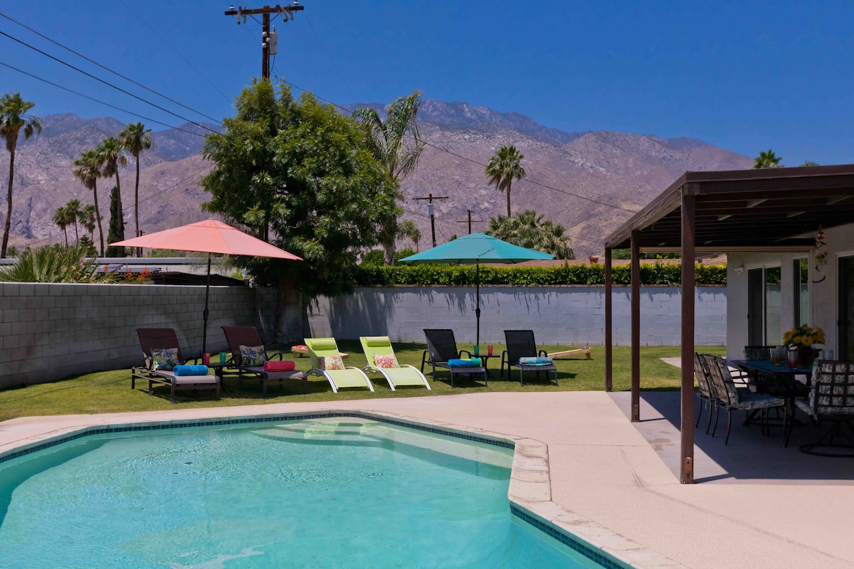 A well-maintained backyard features a heated pool surrounded by comfortable lounge chairs with colorful cushions. Two umbrellas provide shade, while a dining area with a table is positioned under a covered patio. Lush grass and mountain views complete the serene outdoor space.