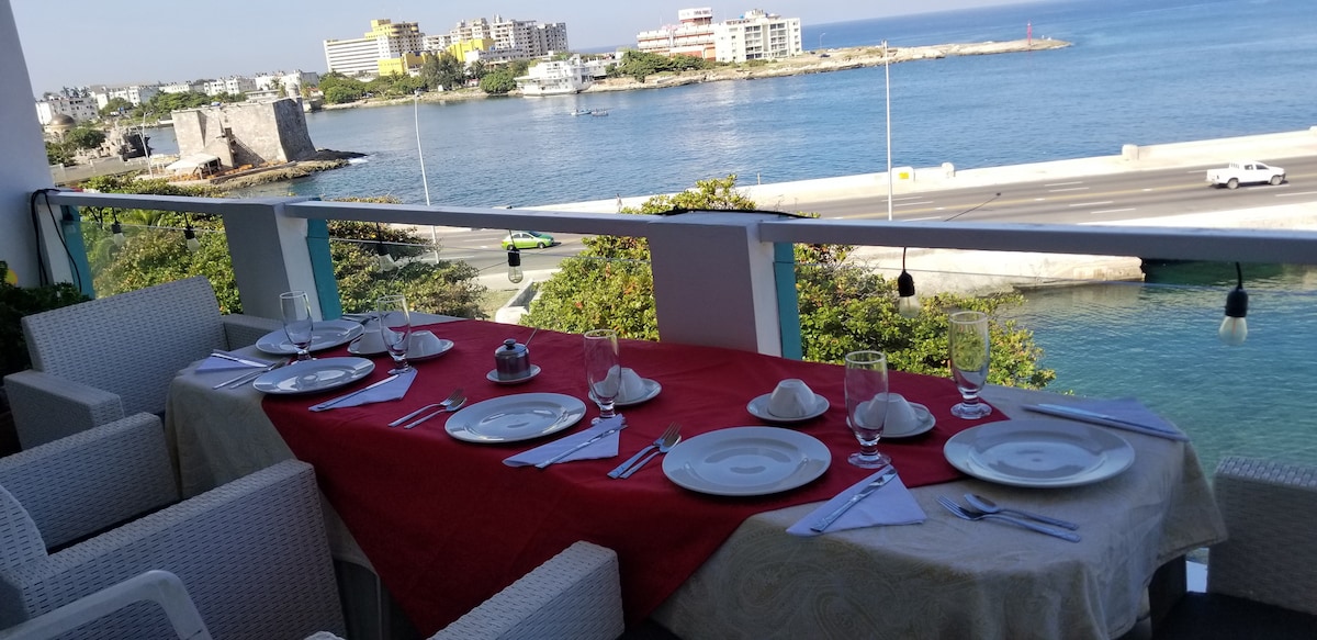 A dining area features a long table draped with a red tablecloth, set with plates and utensils. It overlooks a calm sea, with views of distant buildings and boats. Soft lighting is provided by hanging fixtures along the balcony railing.