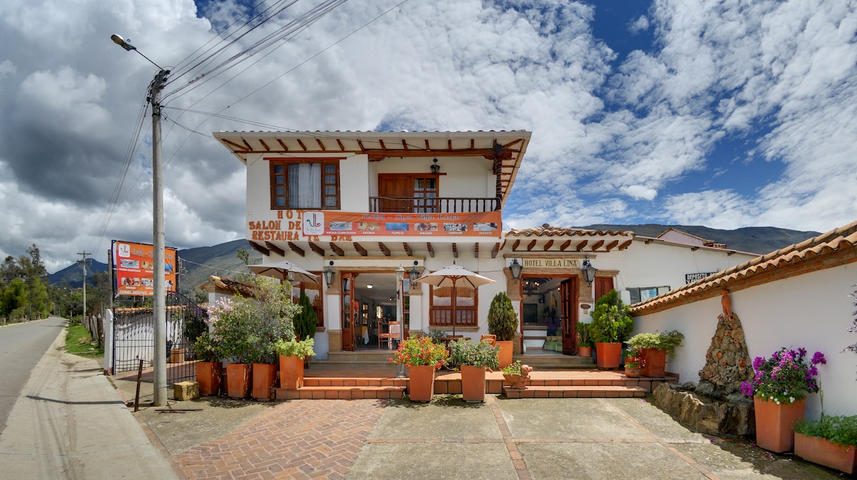 The exterior of the Hotel Spa Villa Lina is displayed, featuring a two-story structure with a traditional design. Large windows are framed by vibrant flowers in terracotta pots. A welcoming entrance is highlighted by a covered porch, with scenic mountains visible in the background.