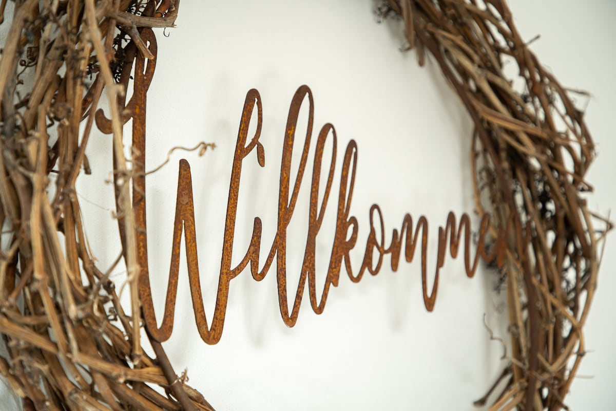 A rustic welcome wreath crafted from intertwined branches adorns a light-colored wall. The metal word 'Willkommen' is displayed in an elegant cursive style, inviting guests into the space.