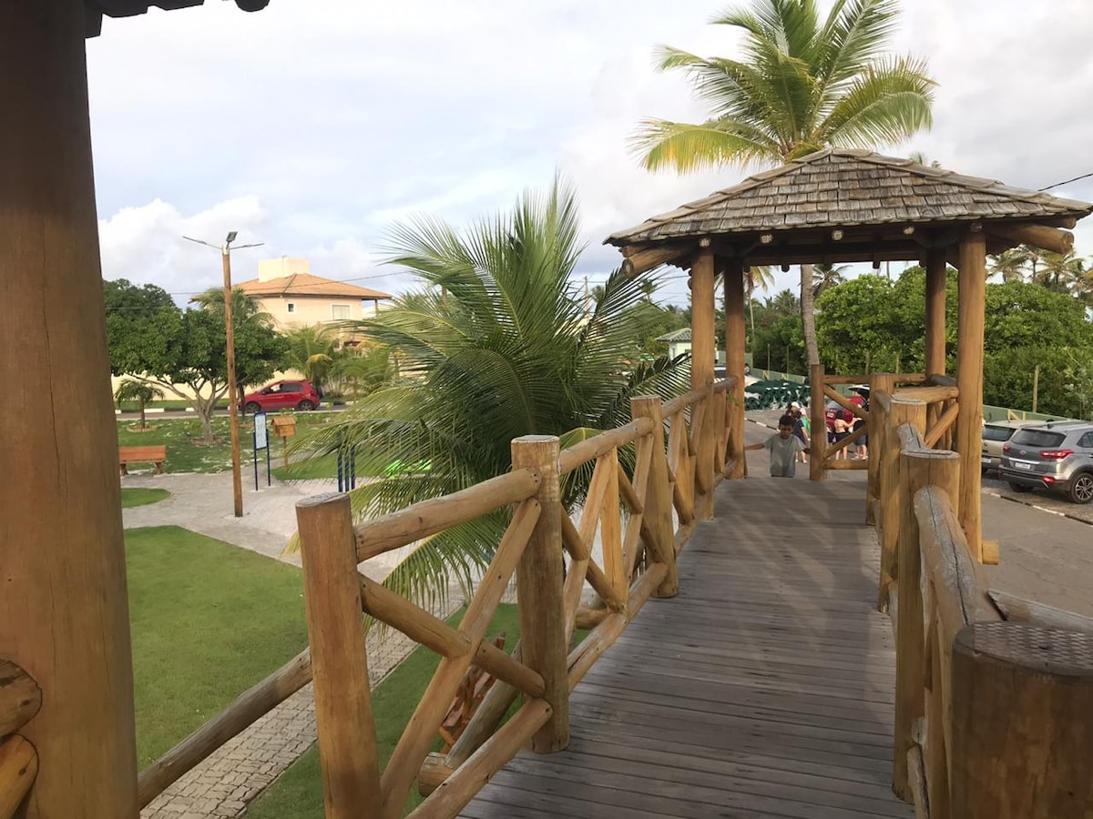 A wooden bridge supported by rustic logs leads to a gazebo, surrounded by lush green palm trees. The walkway provides an inviting view of landscaped grounds, with pathways and parked cars visible in the background, indicating a peaceful communal area.