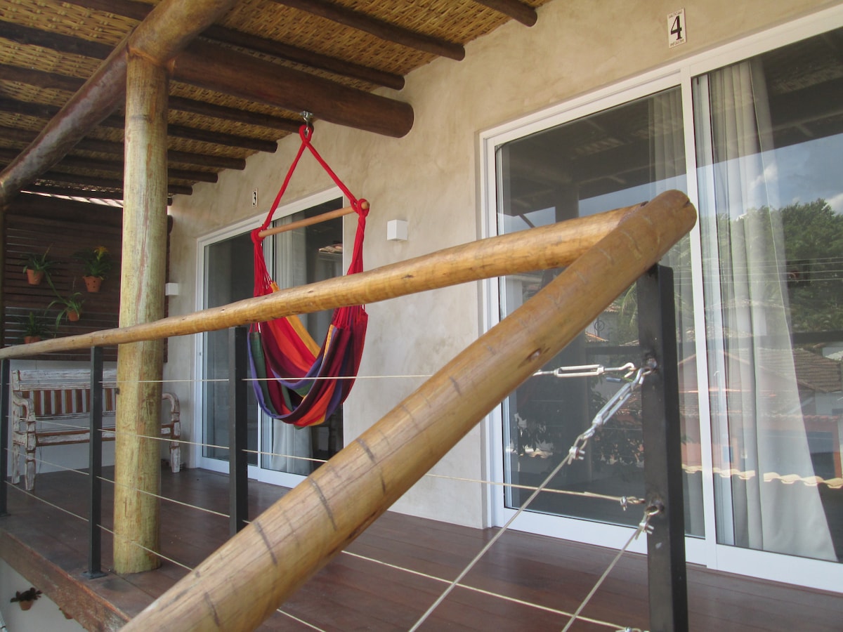 A wooden balcony features a colorful hammock suspended from a rustic frame. Large sliding glass doors provide access to the flats, enhancing the outdoor space. Natural materials and textures create a cozy, welcoming environment, inviting relaxation with views of the surrounding area.