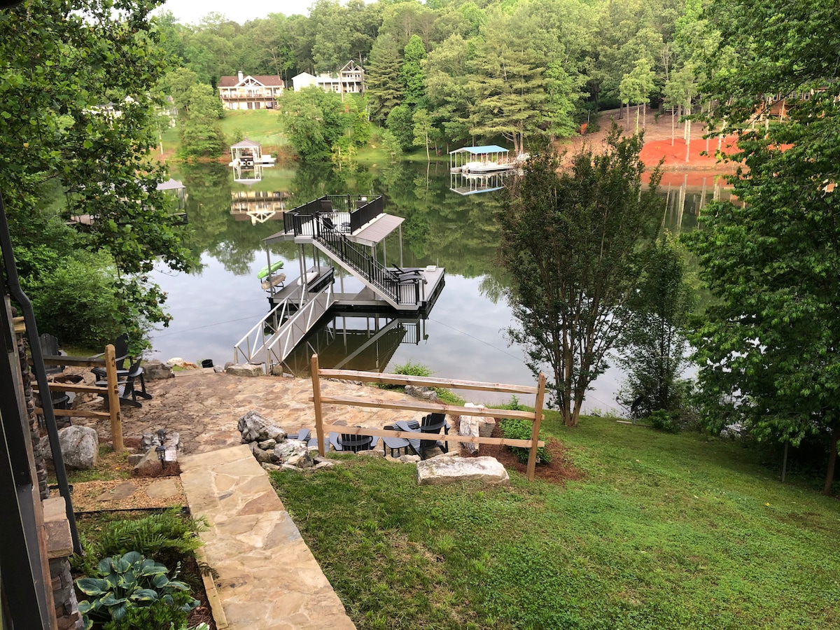 A serene lakeside view displays a wooden dock extending into the calm water. Lush greenery surrounds the area, with a stone pathway leading down to the dock. Adirondack chairs are arranged on the grass, inviting relaxation by the water.