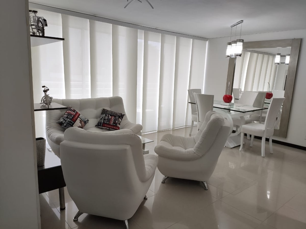 A modern living area features a pair of white armchairs positioned near a sleek glass dining table, accompanied by elegant white chairs. Soft, light-filtering curtains adorn large windows, creating a bright, open atmosphere. Decor elements, such as a decorative bowl and artwork, offer subtle touches of style.
