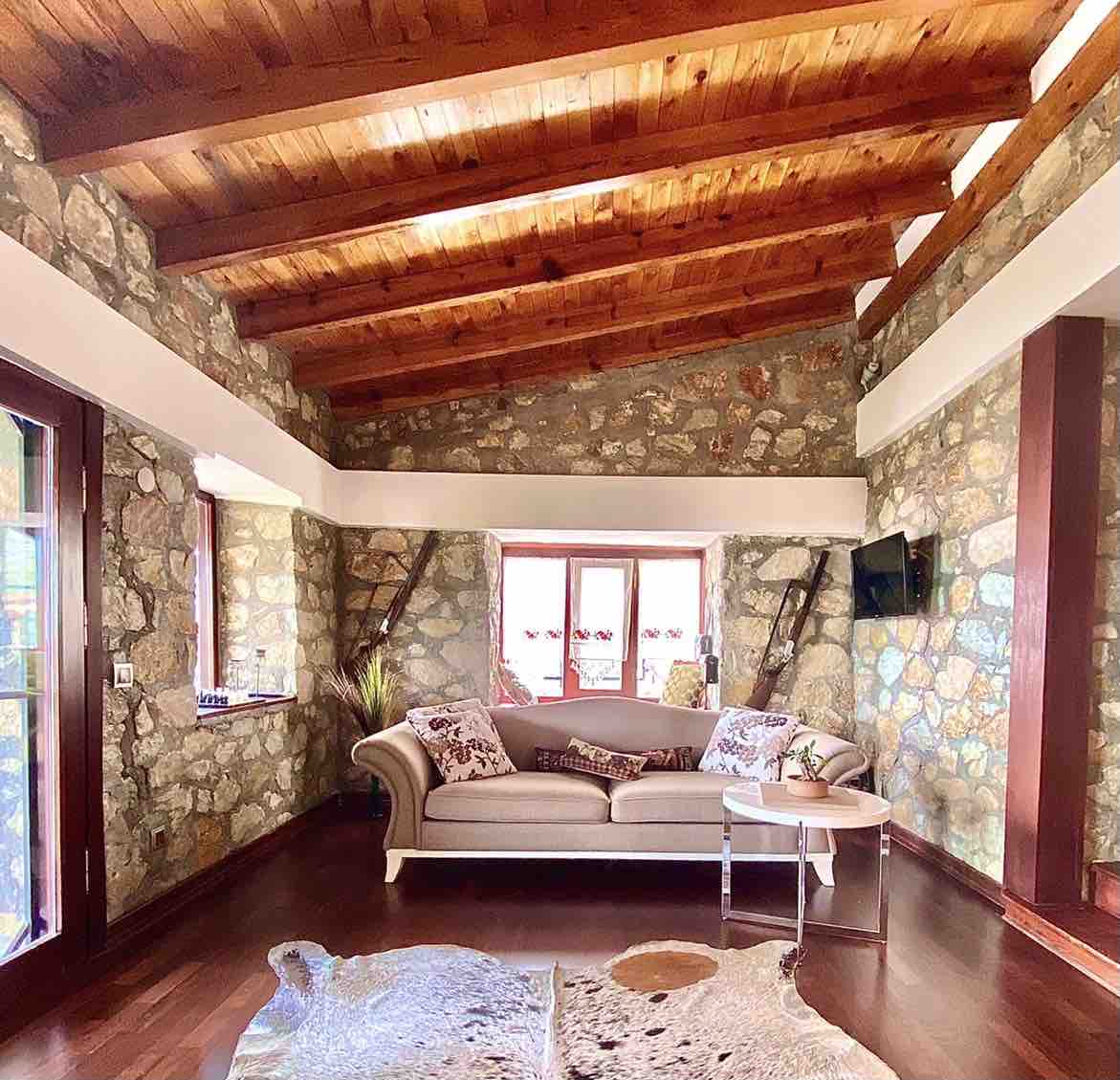 A spacious living area is highlighted by natural stone walls and wooden beams that provide a rustic charm. A comfortable sofa is positioned across from a wall-mounted television. Sunlight streams in through multiple windows, enhancing the inviting atmosphere of the room.