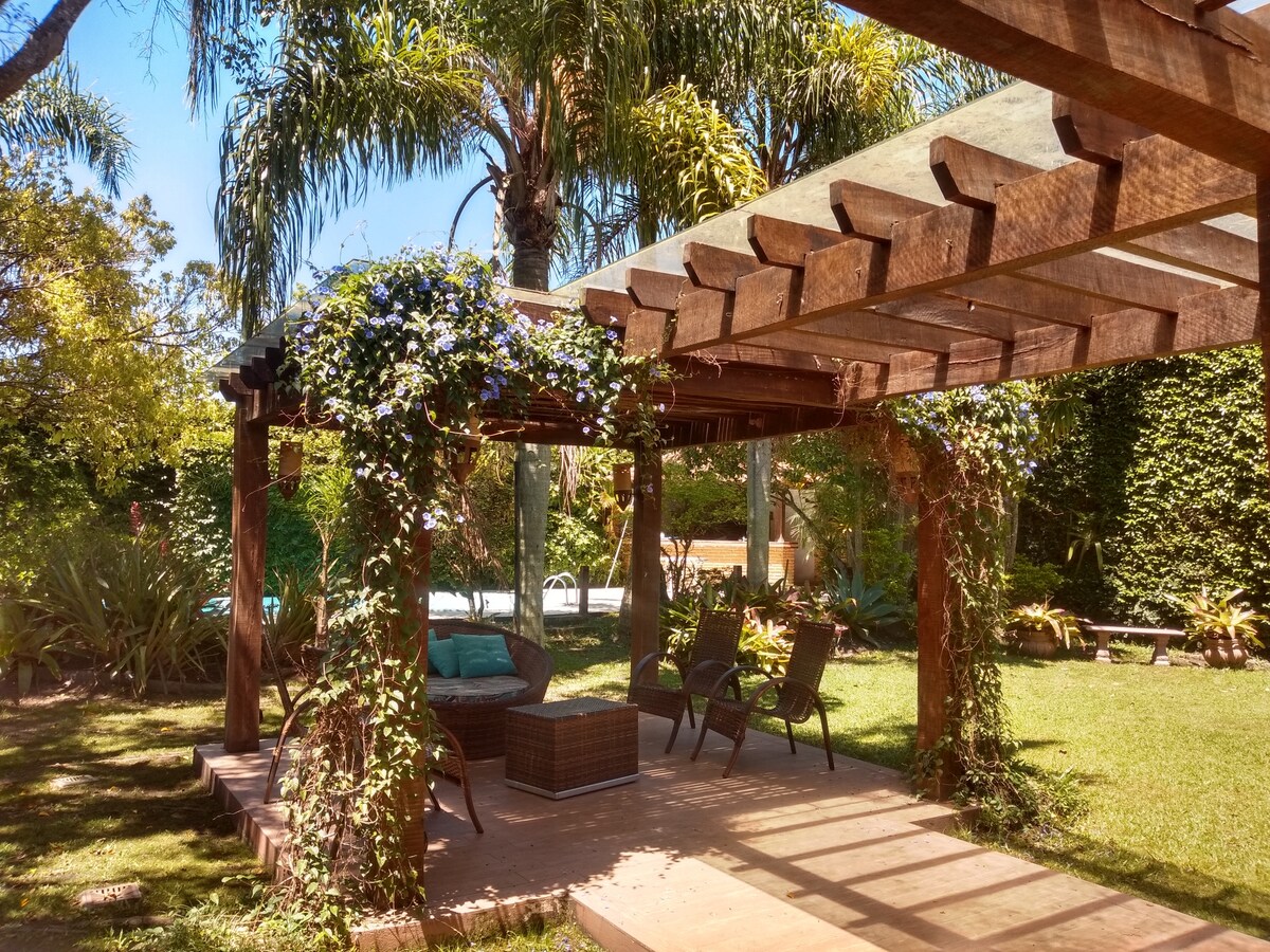 A wooden pergola is surrounded by lush greenery and palm trees, creating a shaded seating area. Comfortable chairs are arranged beneath the structure, with flowering vines climbing along the beams. In the background, a serene pool is visible, enhancing the outdoor space.