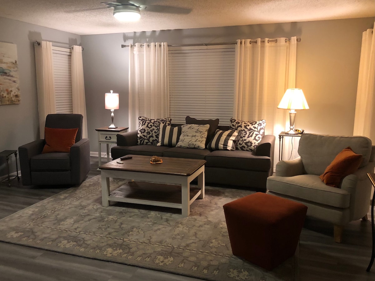 A cozy living room is presented with soft lighting and comfortable seating. The space includes a spacious sofa adorned with patterned cushions, a plush armchair, and a stylish ottoman. A wooden coffee table sits at the center, complemented by decorative accents and light-colored curtains framing the windows.