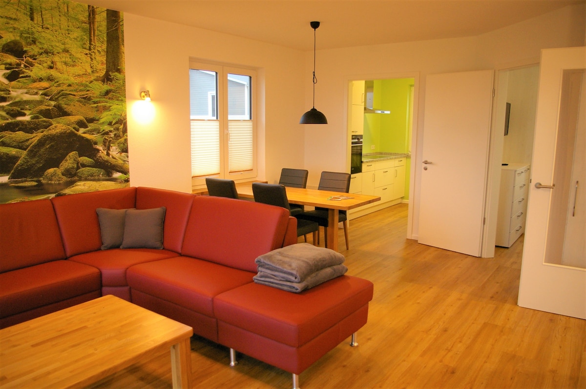 A modern living area features a spacious red sectional sofa with neatly arranged cushions. A wooden coffee table sits in front of the sofa. In the background, a dining table with black chairs and a bright kitchen are visible, complemented by natural light through the windows.