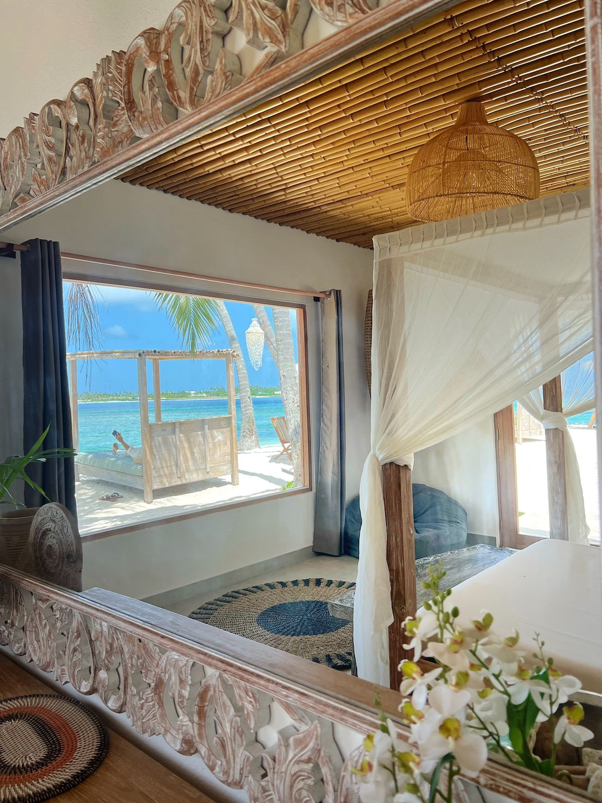 A spacious room showcases a large window framed by tropical foliage, with a view of the turquoise sea. Natural light floods the space, highlighting a cozy bed draped with sheer white curtains. A decorative mirror reflects the bamboo ceiling and surrounding elements, creating an inviting ambiance.