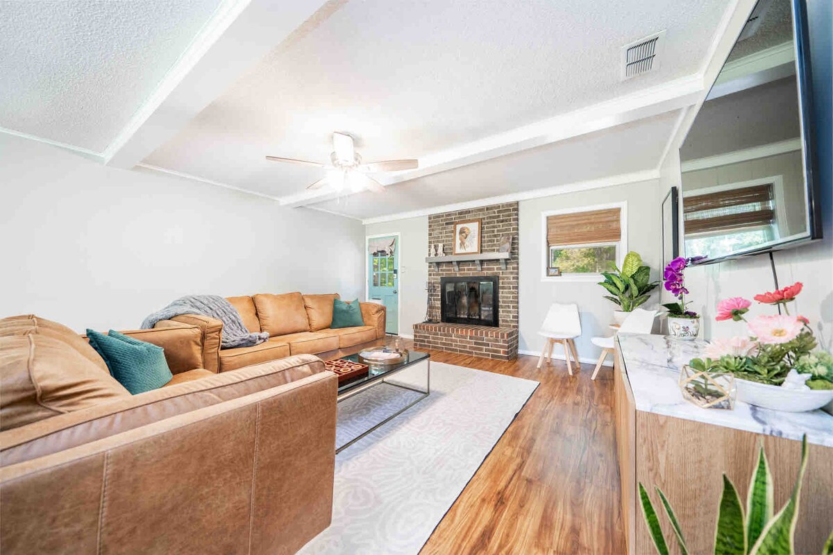 A comfortable living room features a spacious sectional couch and a decorative coffee table. A fireplace is placed against a brick wall, while natural light enters through large windows with wooden blinds. Plants and decorative elements are arranged on a side table.