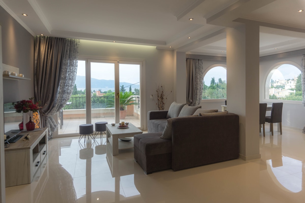 A spacious lounge area is illustrated with modern furnishings including a large sofa, accent chairs, and a coffee table. Large glass doors invite natural light, revealing views of the surrounding greenery and mountains. Elegant curtains frame the windows, enhancing the bright and airy environment.