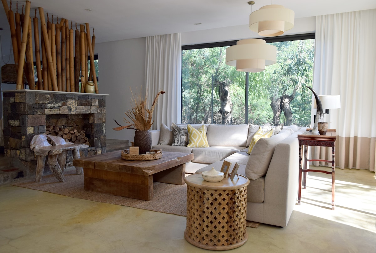 An airy living room features a large L-shaped sofa adorned with cushions, positioned around a wooden coffee table. Natural light floods through expansive windows, offering views of the surrounding greenery. A stone fireplace and decorative accents add warmth to the inviting space.