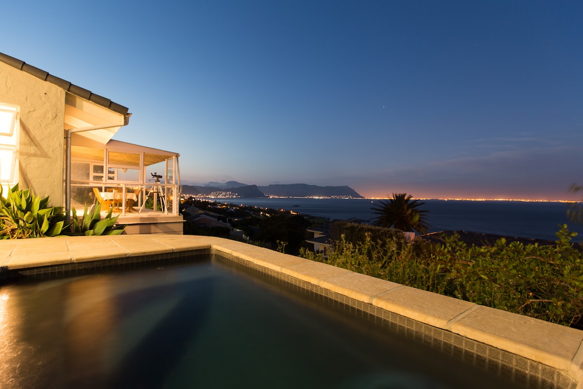 The image captures a tranquil evening view from a villa overlooking False Bay. A modest swimming pool is seen in the foreground, while the coastline and distant mountains stretch across the horizon, illuminated by a soft, natural twilight glow.