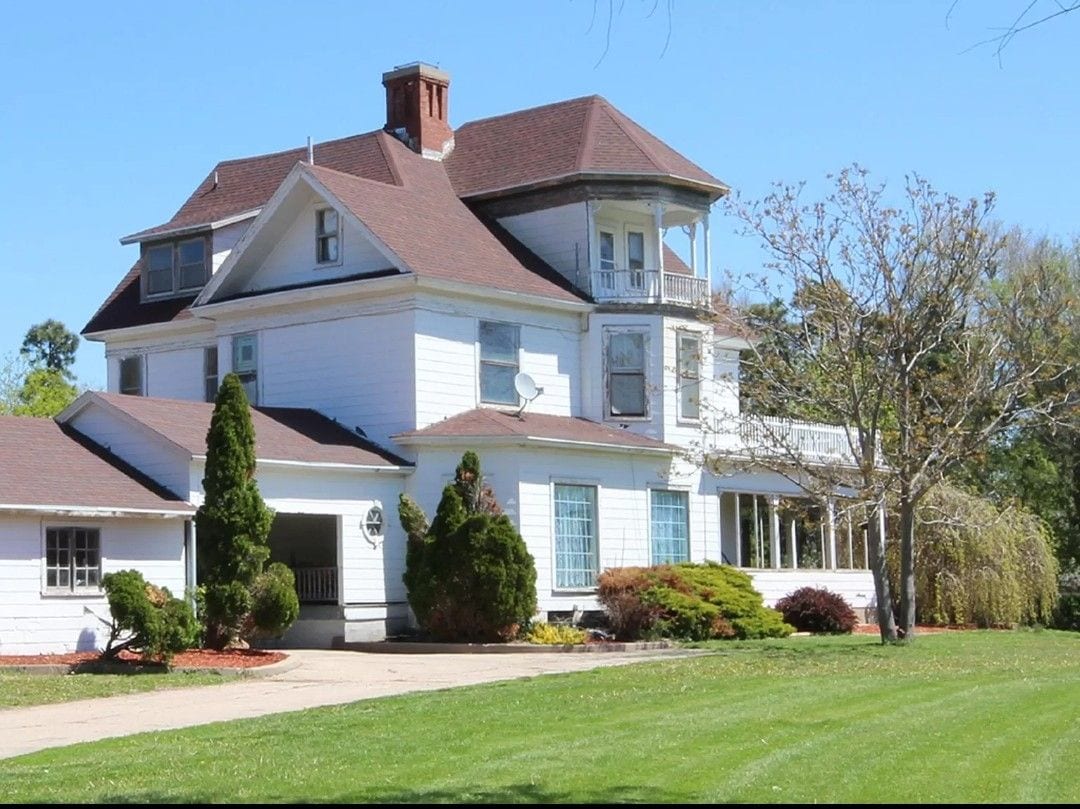 A large, multi-story house sits surrounded by well-maintained greenery. The structure features various architectural details, including large windows and a wraparound porch. The manicured lawn is complemented by trees and shrubs, adding to the home's inviting exterior.
