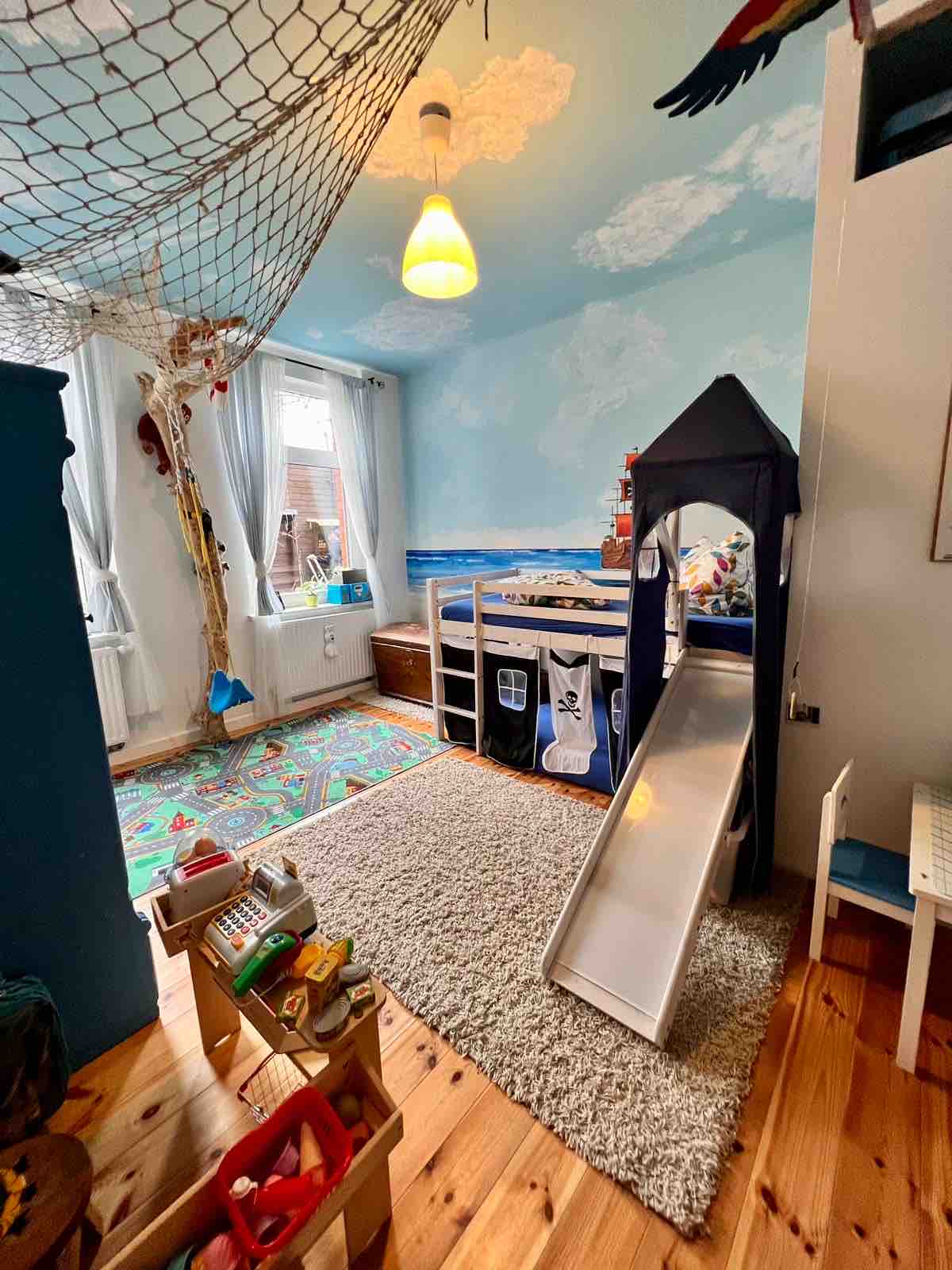 A child-friendly room features wall art of clouds and ocean, with a loft bed and a slide. A colorful rug is placed on the wooden floor, while a small table and chairs are visible. Toys are neatly arranged in a play area, enhancing the playful environment.