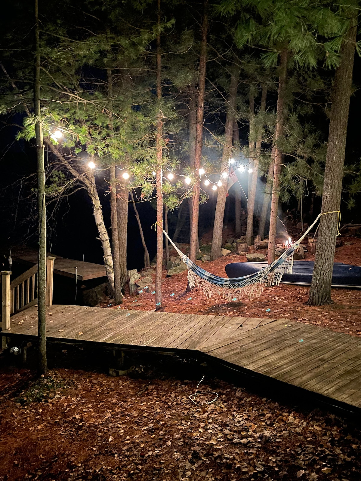 A tranquil lakeside scene is illuminated by string lights strung above a cozy hammock, surrounded by tall trees. A wooden walkway leads to the dock, where additional seating can be seen, providing a relaxing atmosphere for evening gatherings.