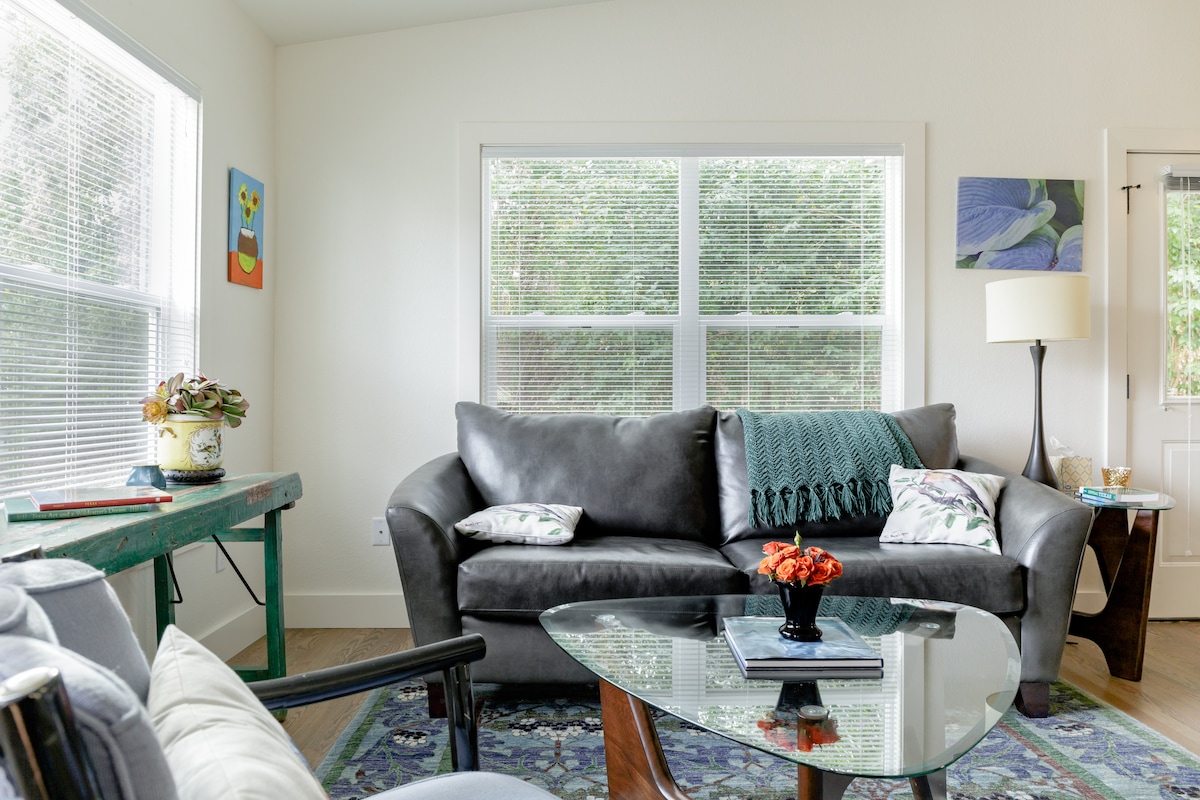 A bright living area features large windows that provide natural light. A dark leather sofa is complemented by decorative pillows. A glass-topped coffee table is positioned at the center, surrounded by an area rug. A small table with a plant adds an inviting touch.