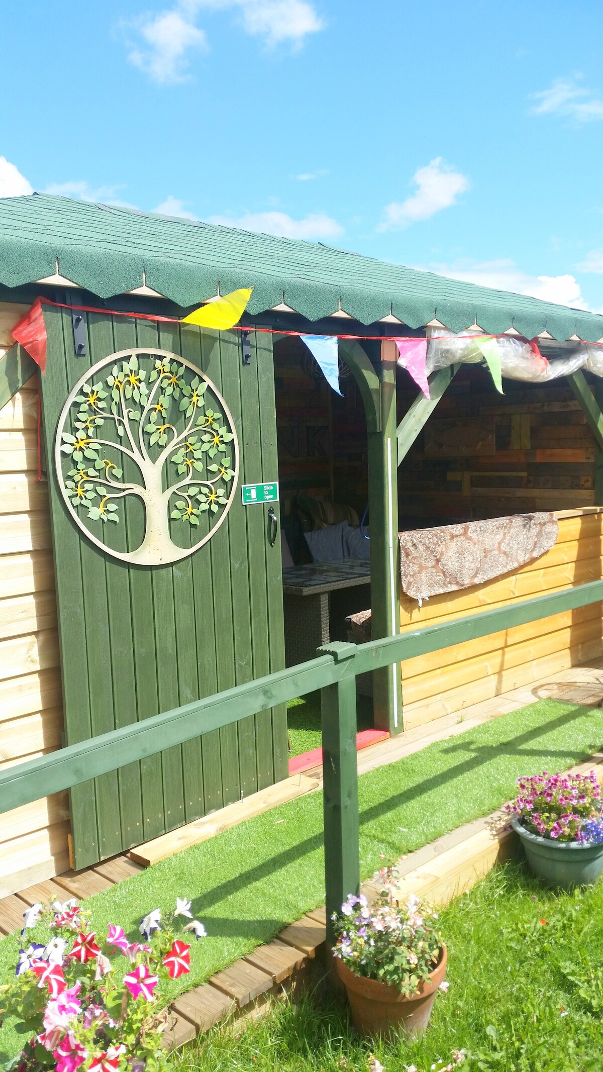 A charming wooden structure features a green door adorned with a tree design. Colorful bunting hangs from the roof, while a well-maintained garden path leads to the entrance. Bright flowers are positioned along the pathway, adding a touch of color to the serene outdoor setting.