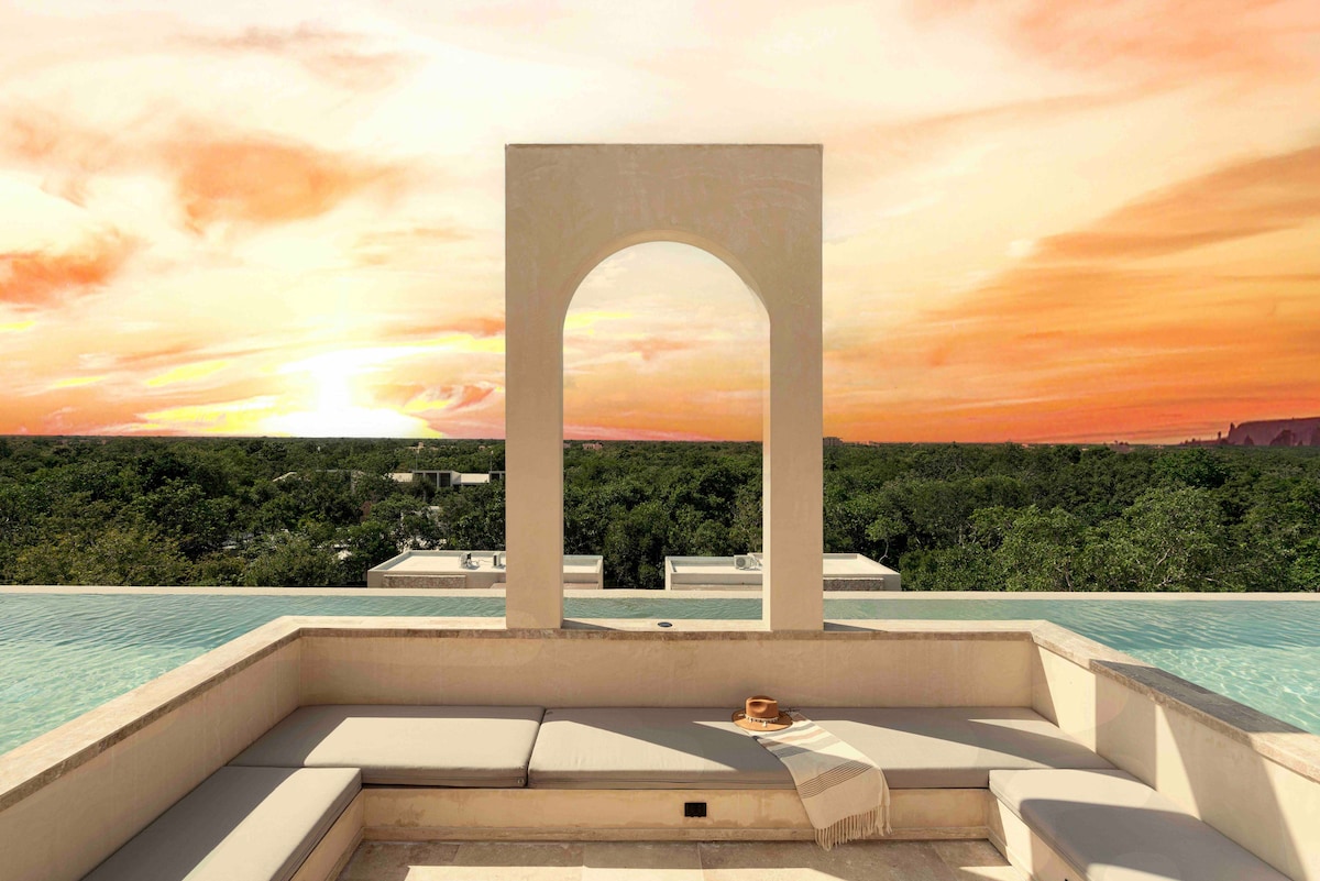 A spacious rooftop terrace features a stylish lounge area with neutral seating positioned beneath an archway. The vibrant sunset casts warm hues over the lush green landscape, creating a serene backdrop for relaxation.