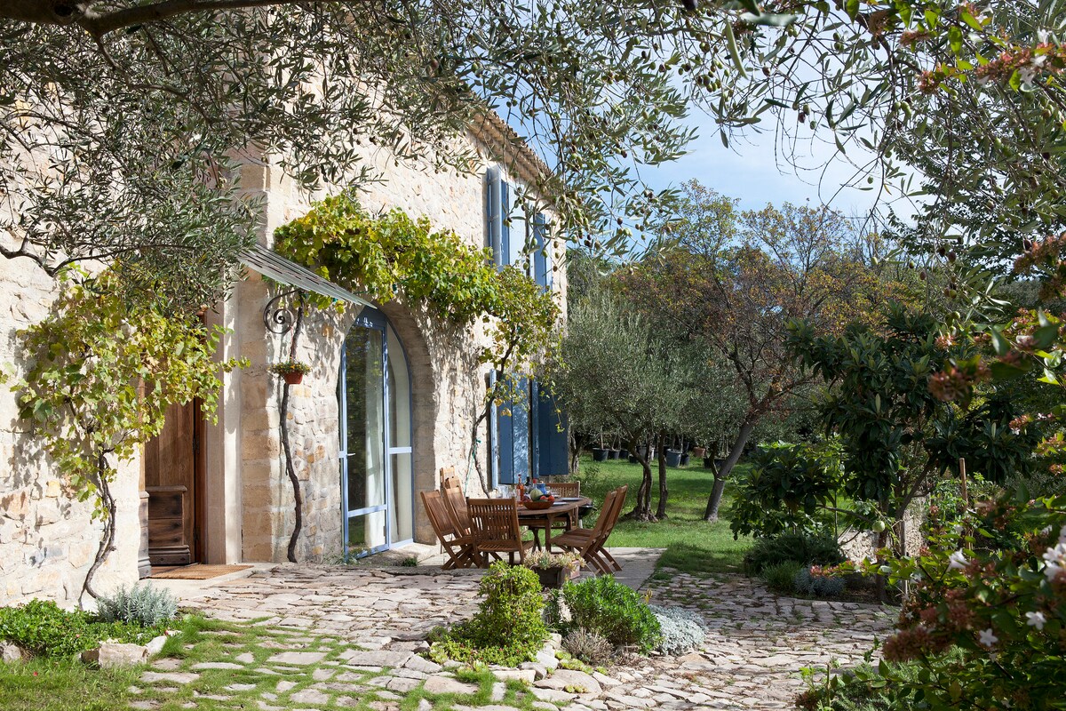 A stone facade of the bastide is framed by lush greenery and Mediterranean plants. A wooden dining table with matching chairs is set on a stone patio, inviting outdoor dining. The entrance features large windows that offer views of the expansive garden.