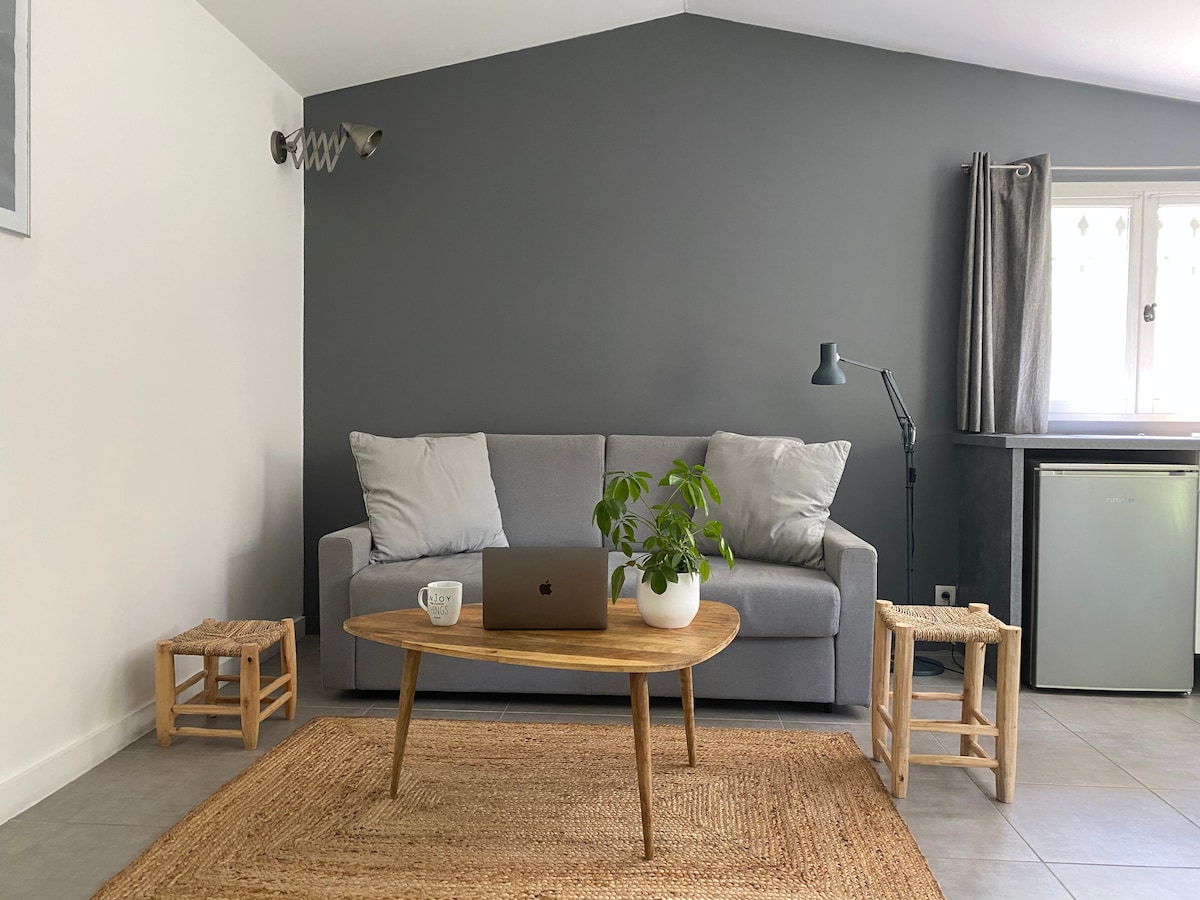 A modern living space features a gray accent wall with a comfortable sofa flanked by two small bamboo stools. A wooden coffee table sits in front of the sofa, accompanied by a laptop and a potted plant. Natural light enters through a window dressed with light curtains.