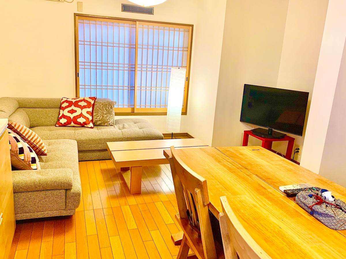 A spacious living area features a large sectional sofa adorned with decorative pillows, complemented by a low coffee table. A wooden dining table with four chairs is positioned nearby, and a television is mounted against the wall, offering comfort and functionality.
