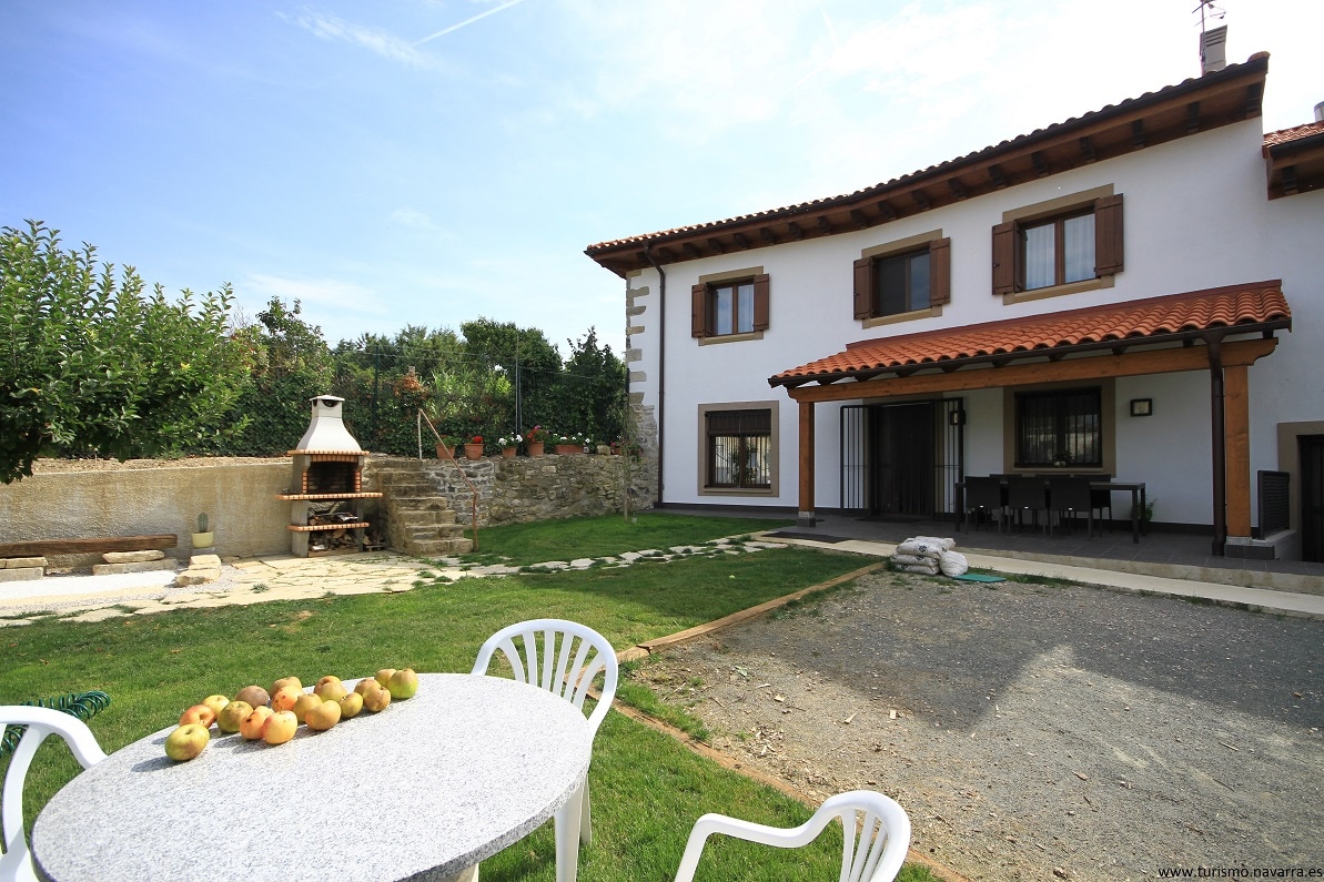 An outdoor area features a round table with white chairs, complemented by a grassy lawn and stone pathway. The house, with a terracotta roof, has large windows and a covered porch. A barbecue grill is present among the greenery, offering a tranquil setting.