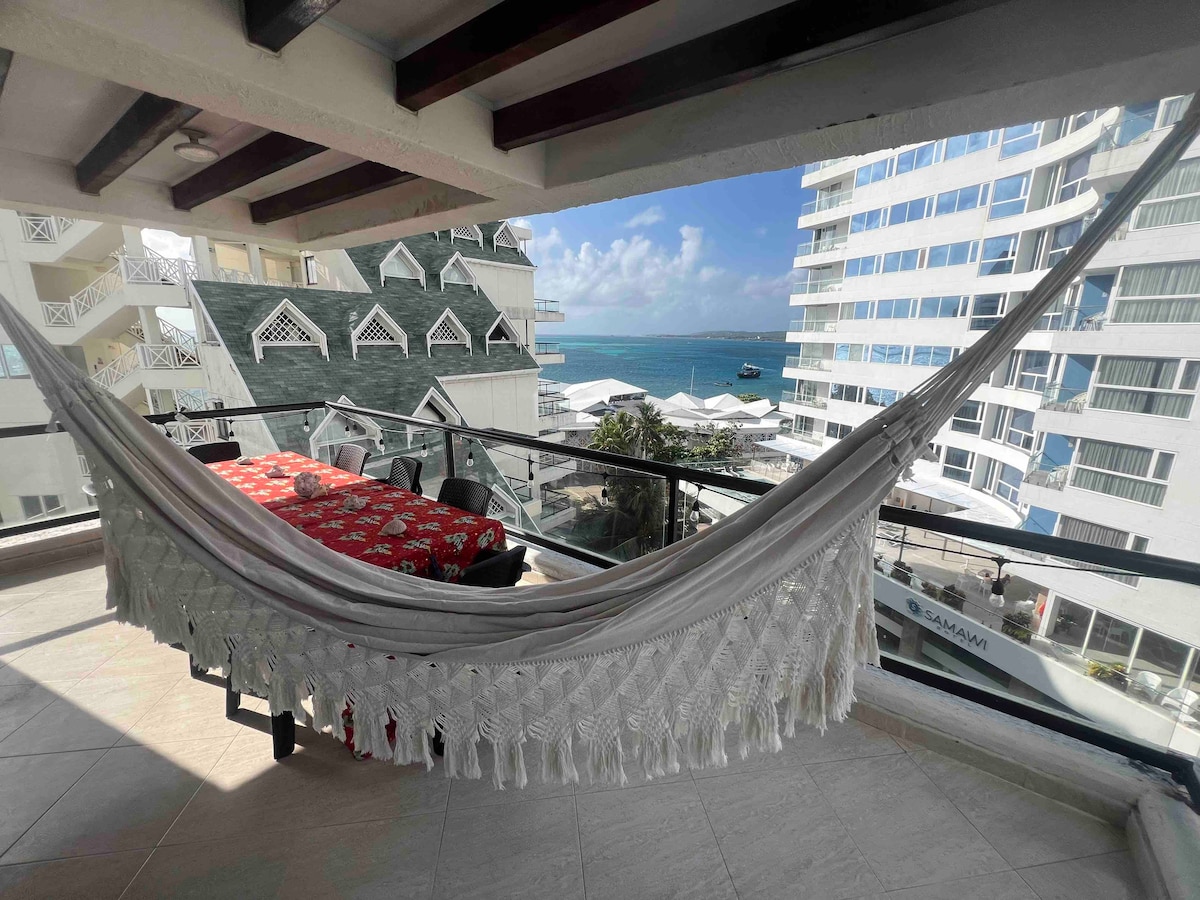 A balcony features a macramé hammock draped elegantly, alongside a dining table covered with a patterned tablecloth. Glass railings provide a clear view of the ocean and surrounding buildings, with bright skies enhancing the overall ambiance.