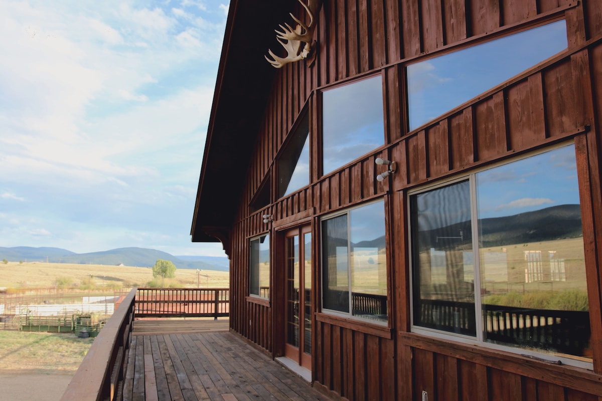The exterior of the Lodge features a wooden façade with large windows, reflecting the surrounding landscape. A spacious deck extends from the entrance, providing unobstructed views of the ranch and distant mountains. Antlers are mounted above the entryway, adding a rustic touch.
