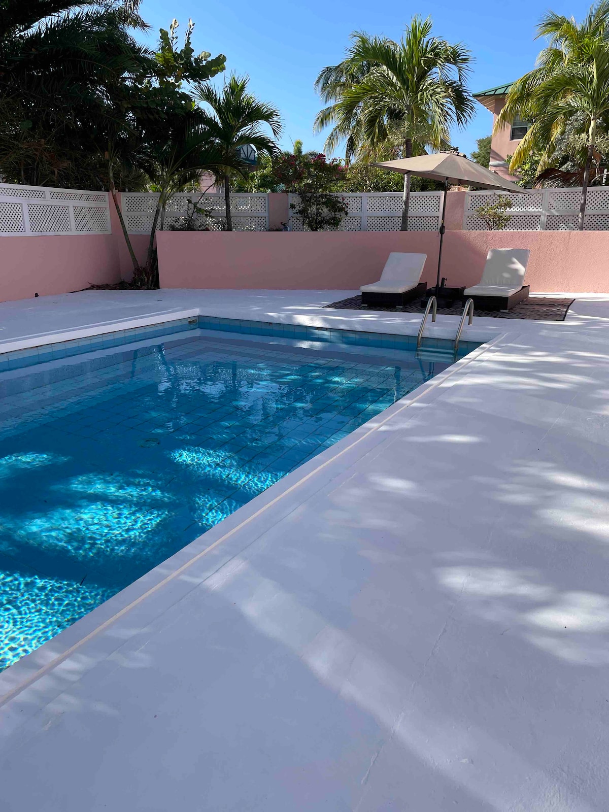 A private swimming pool is surrounded by a white deck and pink privacy walls. Sun loungers are positioned nearby, and tropical plants frame the area, offering a serene setting. The clear water reflects sunlight, creating a calming atmosphere for relaxation.