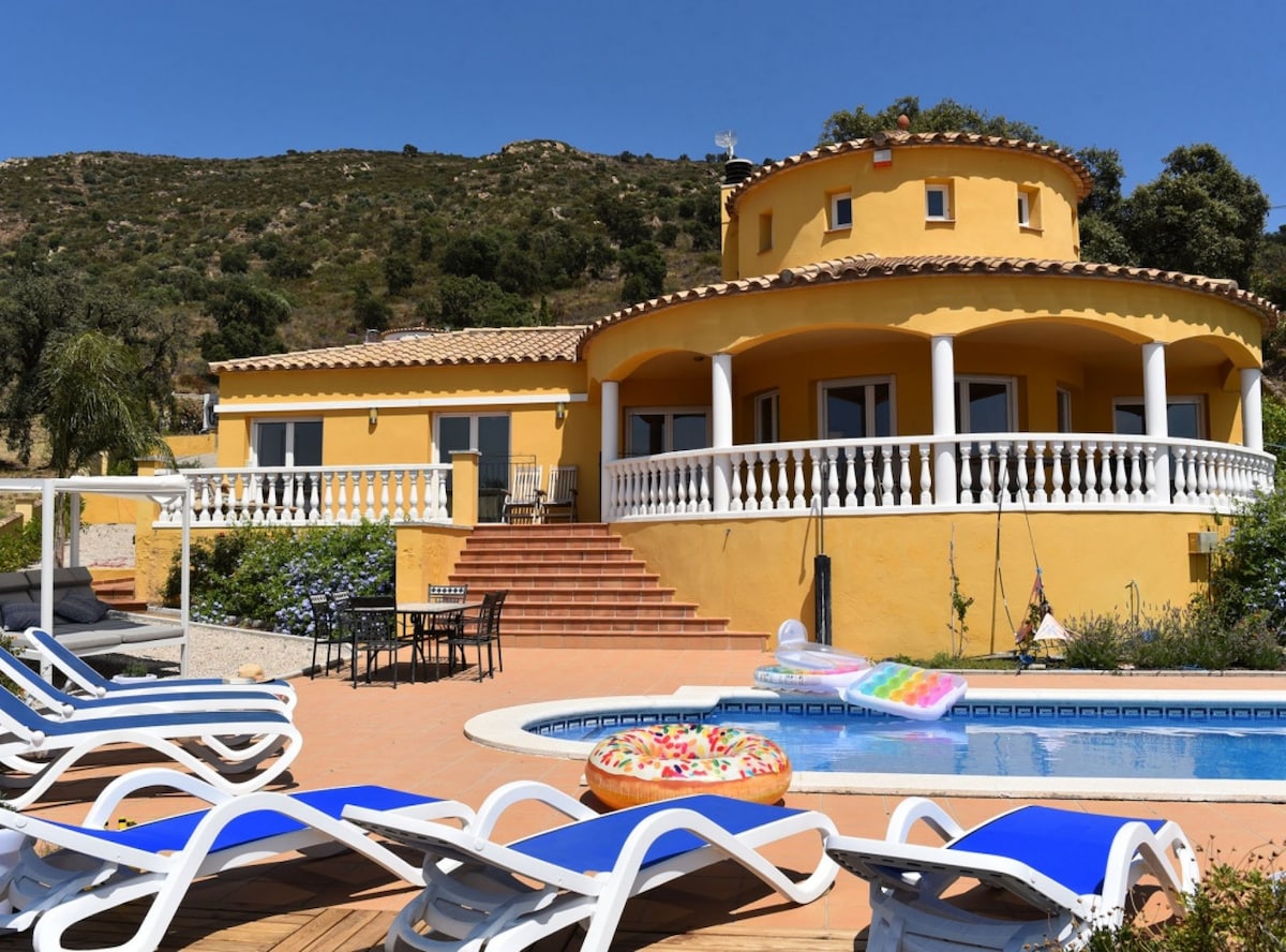 A spacious villa is seen featuring a circular design with a vibrant yellow exterior. The terrace is equipped with loungers and a table, accented by steps leading to the pool area. Lush greenery and hills provide a scenic backdrop to the outdoor space.