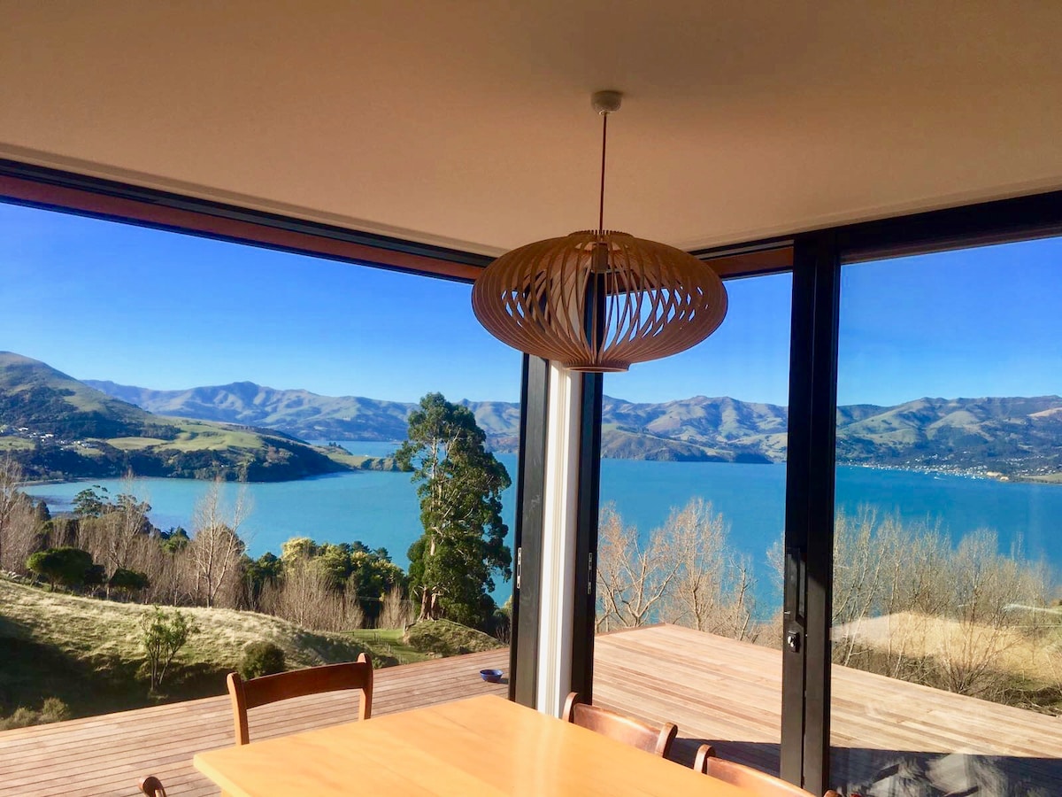A spacious dining area features a wooden table surrounded by chairs, with large glass windows showcasing unobstructed views of Akaroa Harbour and rolling hills in the background. Natural light fills the room, highlighting the sleek design and pendant light overhead.
