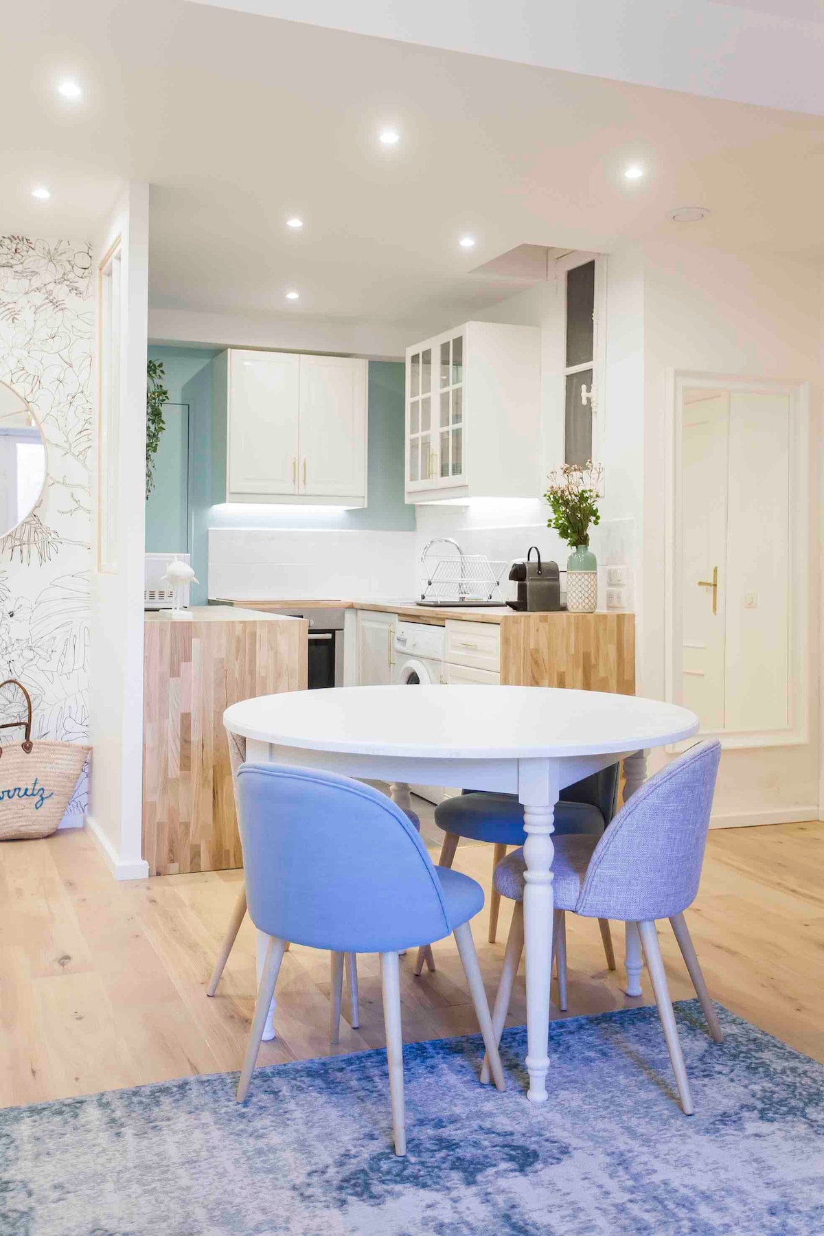 A bright kitchen area features a round dining table with four chairs, surrounded by modern cabinetry. Light wood flooring complements the blue and white kitchen design, while natural light highlights the space. A partial view of appliances, including a washing machine and sink, is visible.