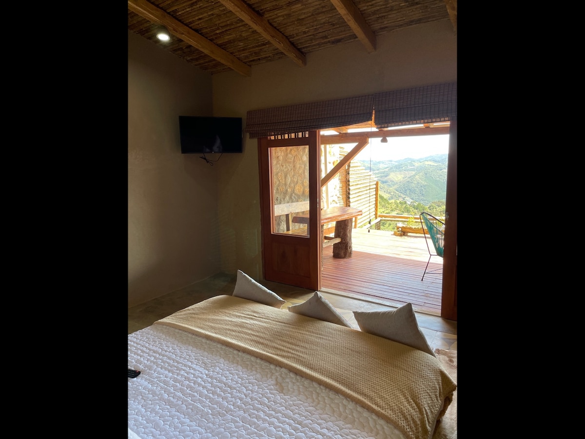 A spacious sleeping area is presented with a neutral-colored blanket laid over the bed. Natural light fills the room from a large glass door that opens to a wooden deck, revealing scenic mountain views. A television is mounted on the wall for convenience.
