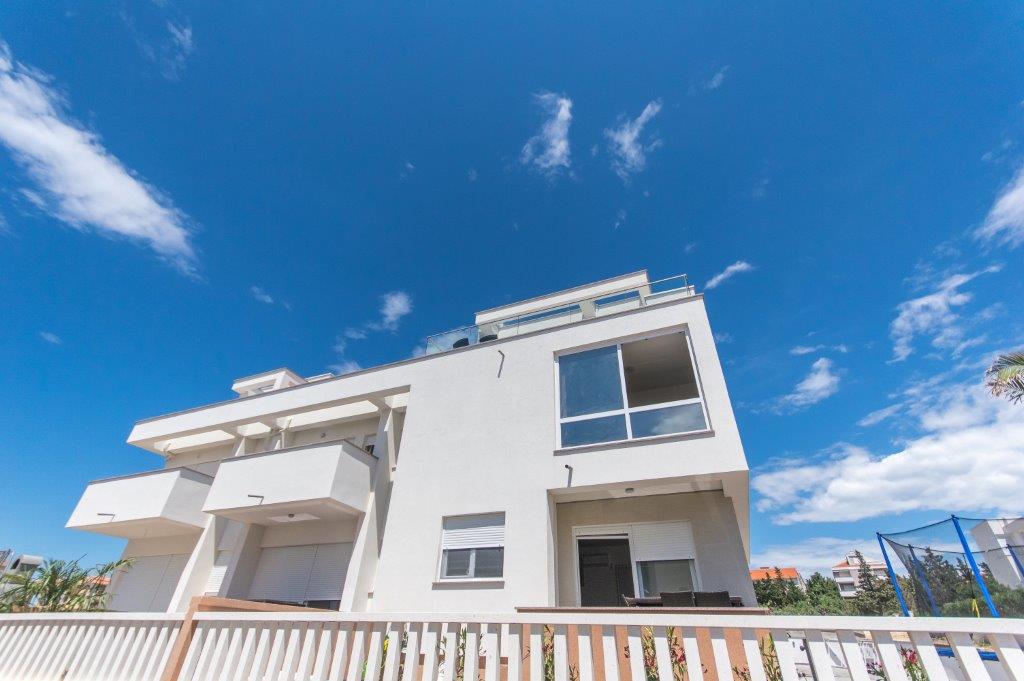 The three-story building is set against a clear blue sky, showcasing a contemporary design. Large windows and balconies create an open feeling, while a white facade adds to the modern aesthetic. A wooden fence surrounds the front, enhancing its inviting appearance.