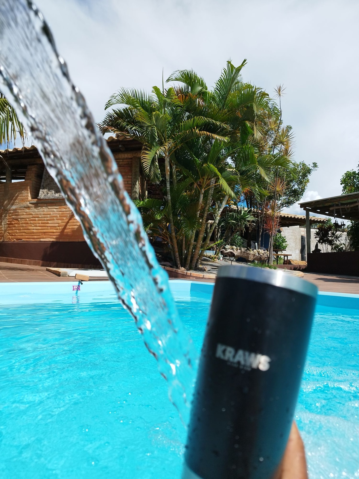 A close-up of a black insulated cup is held by a hand at the edge of a turquoise pool. Water flows from a nearby fountain, creating ripples in the pool's surface. Lush greenery and palm trees are visible in the background, enhancing the serene atmosphere.